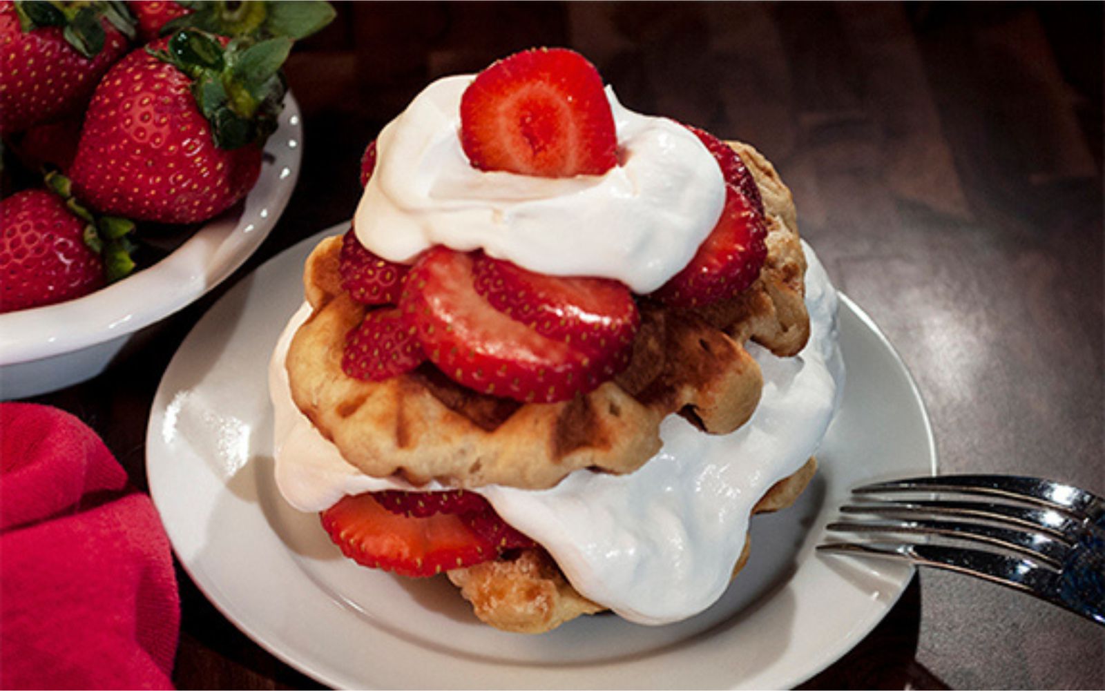 Liège waffles layered with sliced strawberries and whipped cream, with a bowl of strawberries behind